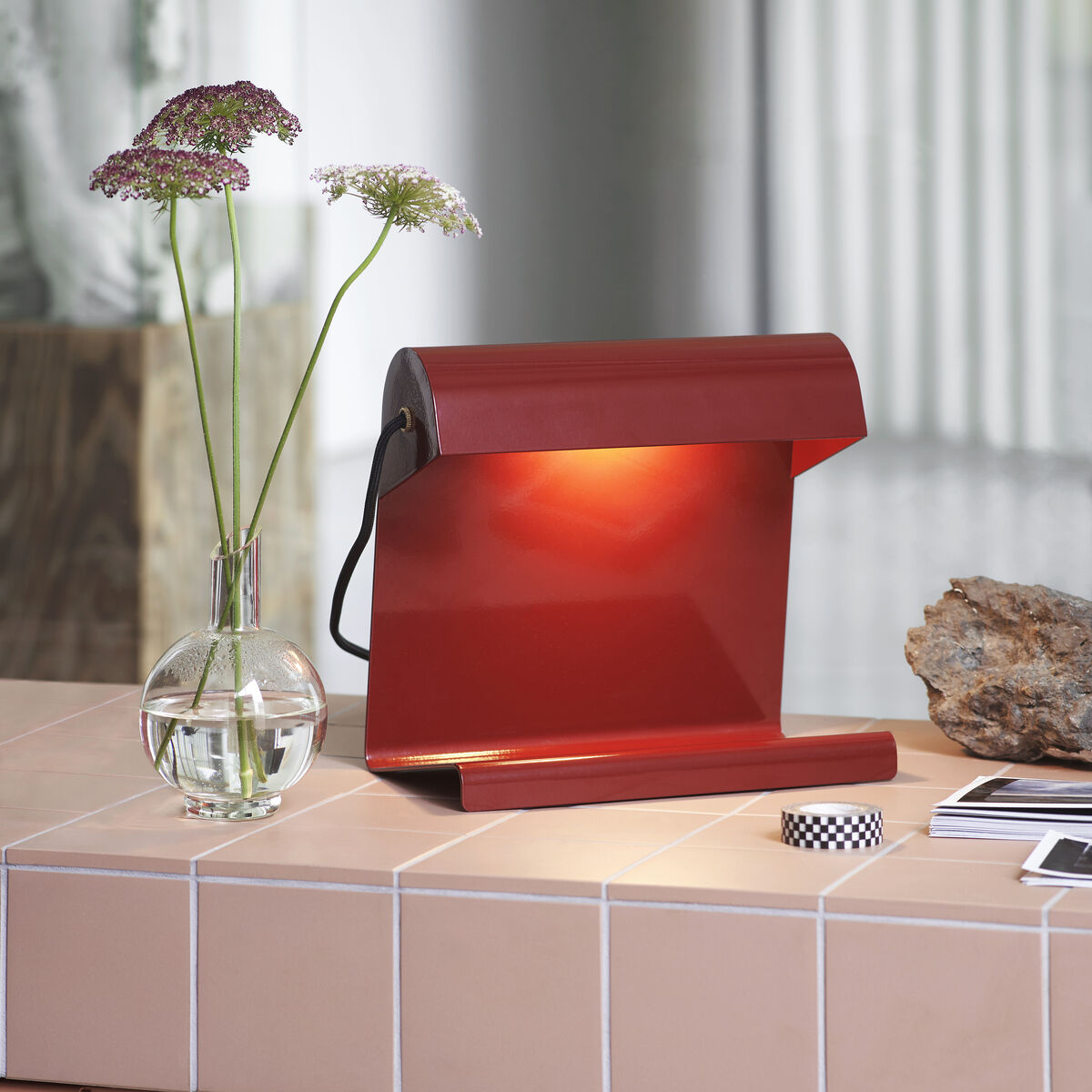 Eine moderne rote Tischlampe »Lampe de Bureau« steht auf einer hellen beige gefliesten Oberfläche neben einer kleinen Vase mit Blumen, einem dekorativen Stein und Bürozubehör. Im Hintergrund sind Strukturen aus Glas und Holz erkennbar.