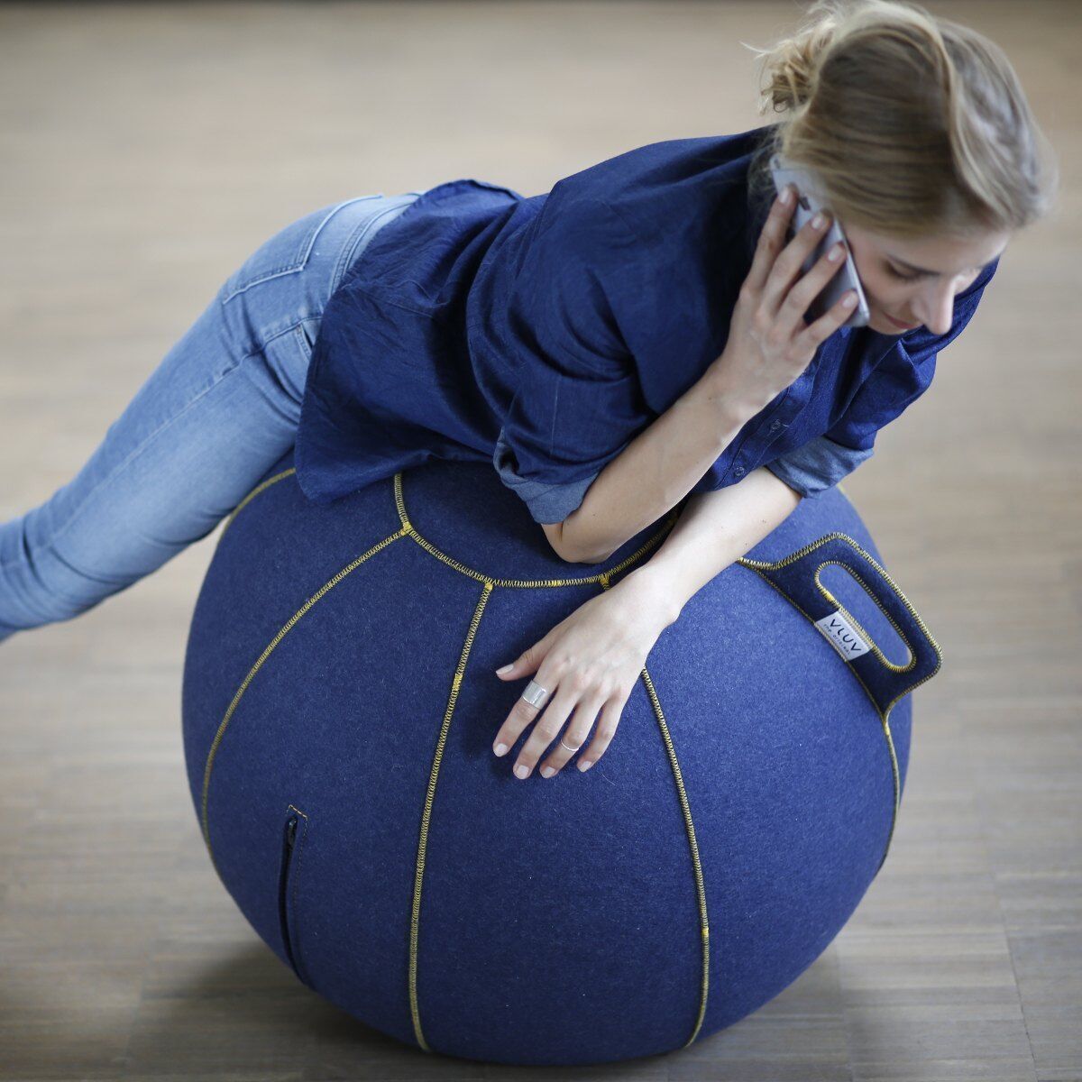 Eine Frau lehnt auf einem Sitzball »Vluv Velt«, telefoniert und trägt eine blaue Jeans und ein blaues Hemd. Der Sitzball ist blau mit gelben Nähten und hat einen Griff. Der Boden ist holzfarben.