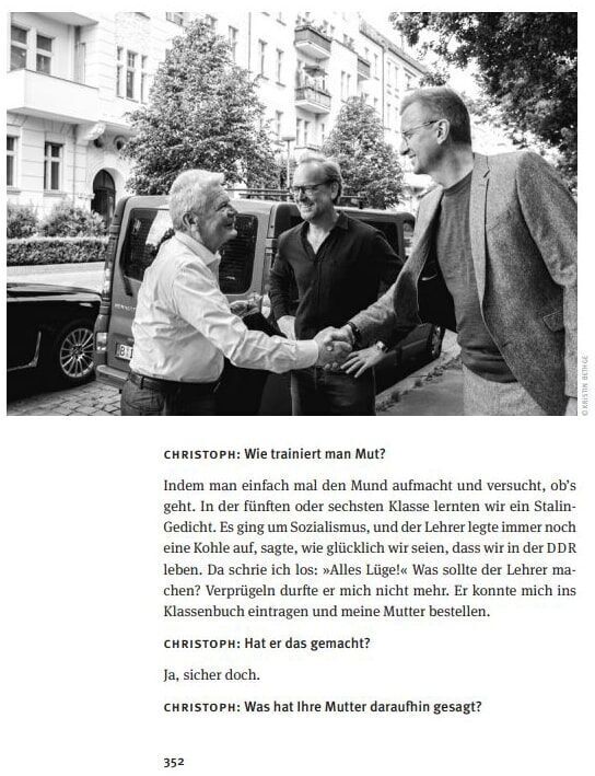 Ein schwarz-weiß Foto aus dem Buch 'Alles Gesagt? Buch zum Podcast', zeigt mehrere Personen im Gespräch auf einer Straßenszene. Darunter ist ein Textdialog in deutscher Sprache abgedruckt.