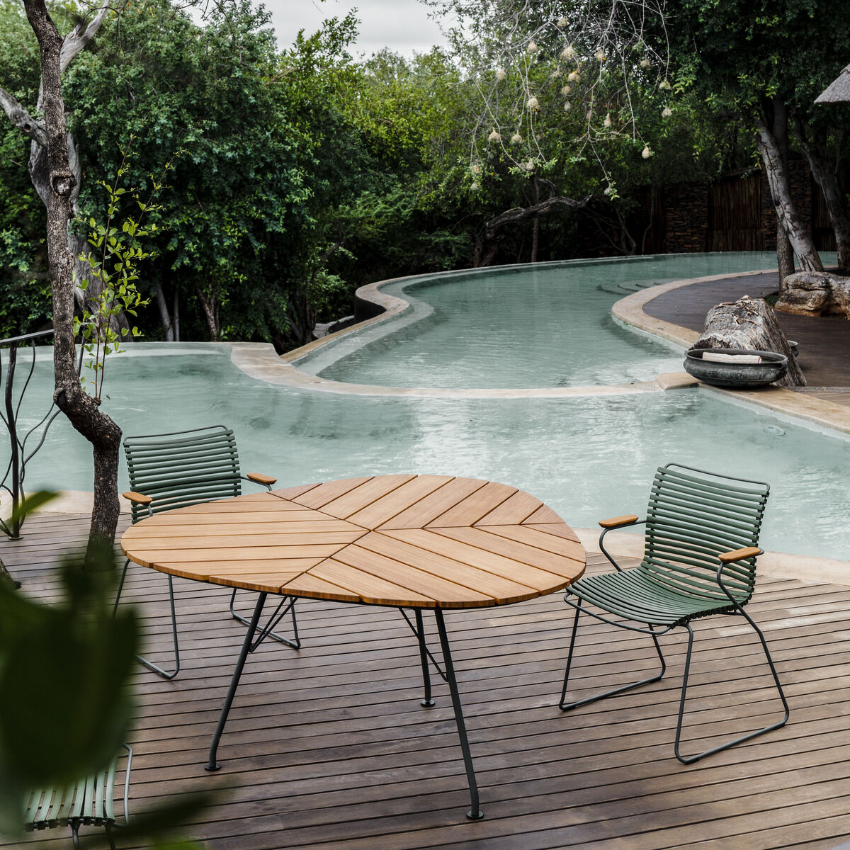 Ein moderner Gartentisch mit Holztischplatte heben sich auf einer Terrasse am Pool hervor, umgeben von grünen Bäumen und mit zwei grünen Metallstühlen.