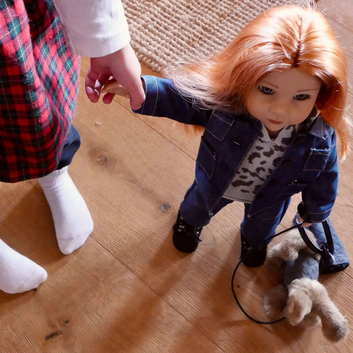 Ein Kind hält die Hand der Stehpuppe »Hannah und ihr Hund Polly« von Götz Puppen. Die Puppe trägt Jeanskleidung, hat lange rotblonde Haare und steht neben einem kleinen Stoffhund auf Holzfußboden. Teil eines Teppichs sichtbar.