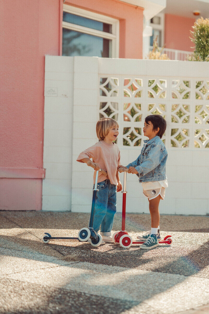 Zwei Kinder fahren auf farbenfrohen Scootern vor einem pastellfarbenen Haus. Einer trägt einen rosa Pullover und Jeans, der andere eine Jeansjacke und weiße Shorts. Im Hintergrund sind weiße und rosa Wände zu sehen.