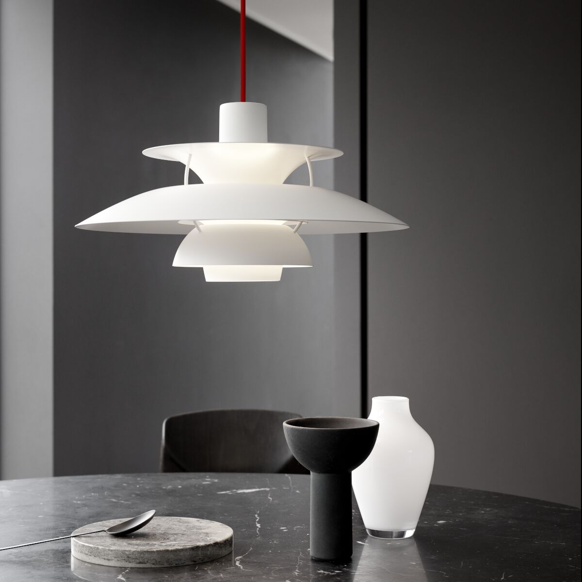 Moderne weiße Pendelleuchte der ZEIT-Sonderedition PH5 Monochrome mit rotem Kabel, stilvoll inszeniert über einem dunklen Marmortisch mit minimalistischen Dekorationen vor einer grauen Wand.