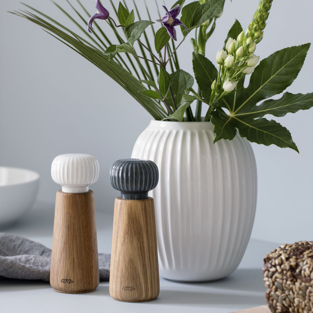 Eine Gewürzmühle »Hammershøi« mit einem mix aus Holz und Porzellan, steht auf einer Küchenarbeitsplatte neben einer weißen Vase mit grünen Pflanzen. Daneben liegt ein graues Tuch, und im Vordergrund liegt ein Stück Körnerbrot.