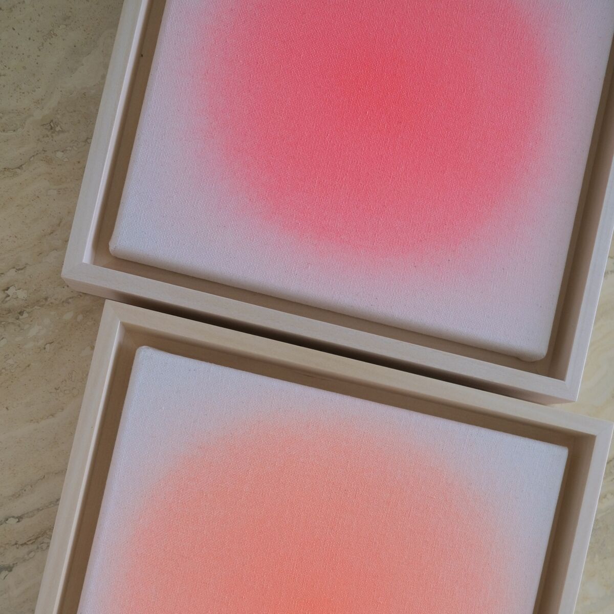 Zwei quadratische Leinwandbilder in einem schlichten Holzrahmen, die jeweils einen zarten Farbverlauf von Weiß zu intensivem Rot und Rosa zeigen. Auf einer strukturierten, beigen Oberfläche platziert. Die harmonische Komposition betont Einfachheit und moderne Ästhetik.