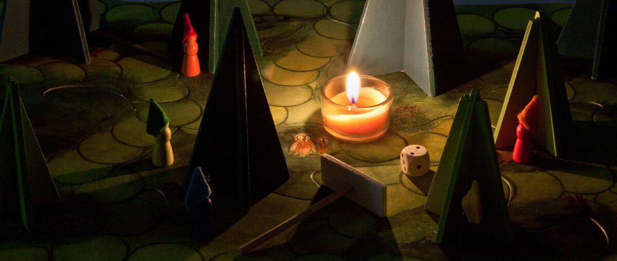 Das Bild zeigt das Brettspiel 'Waldschattenspiel' mit einer brennenden Kerze in der Mitte. Auf dem Spielfeld befinden sich verschiedene grüne Papierbäume, bunte Holzfiguren und ein Würfel. Das Spielbrett zeigt eine dichte Waldlandschaft.