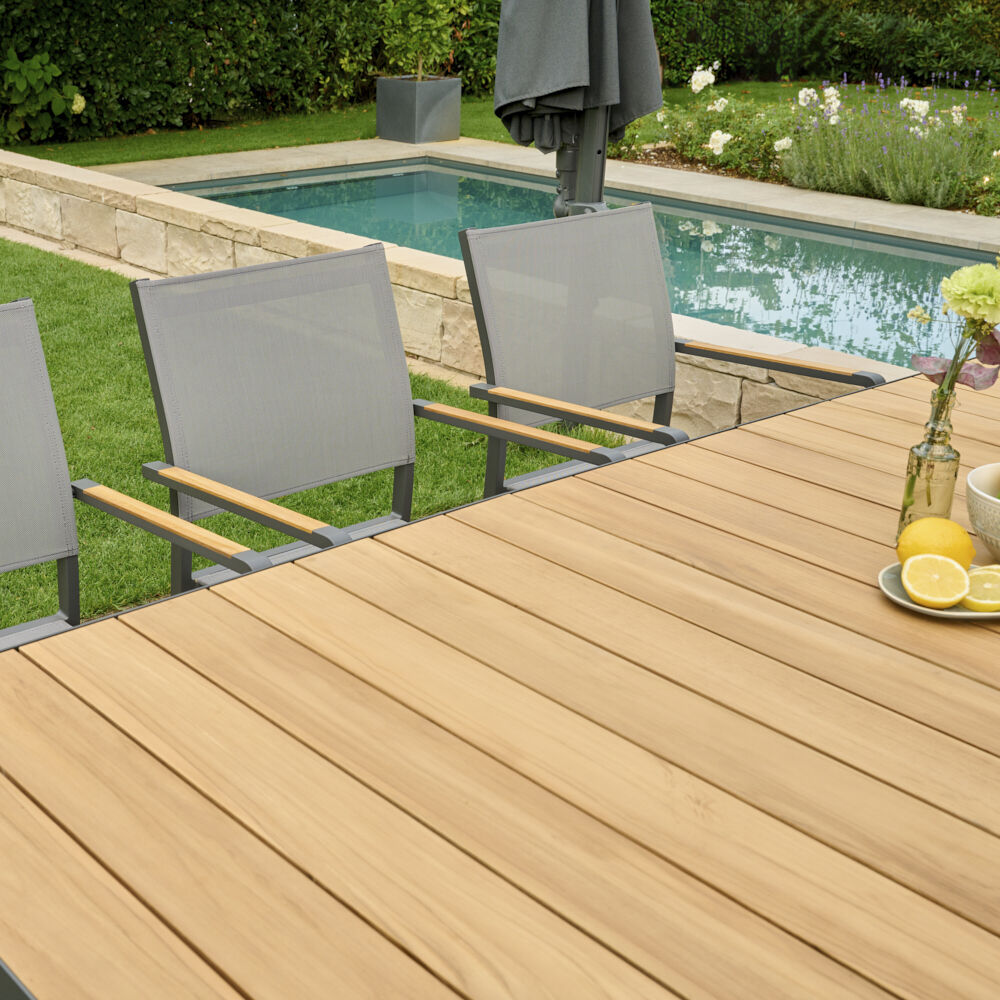 Moderner Ausziehtisch »Sincro« auf einer Terrasse mit Holzoptik und grauen Gartenstühlen, umgeben von Gartenpflanzen und einem kleinen Wasserbecken.