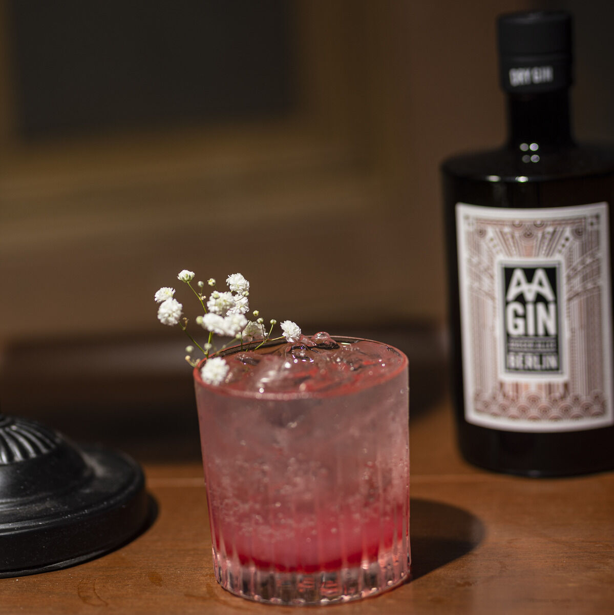 Eine Flasche »AAGIN« Edition 1963 Dry Gin steht auf einem Holztisch. Daneben befindet sich ein Glas mit einem rosa Cocktail und Eiswürfeln, dekoriert mit einer Blume.