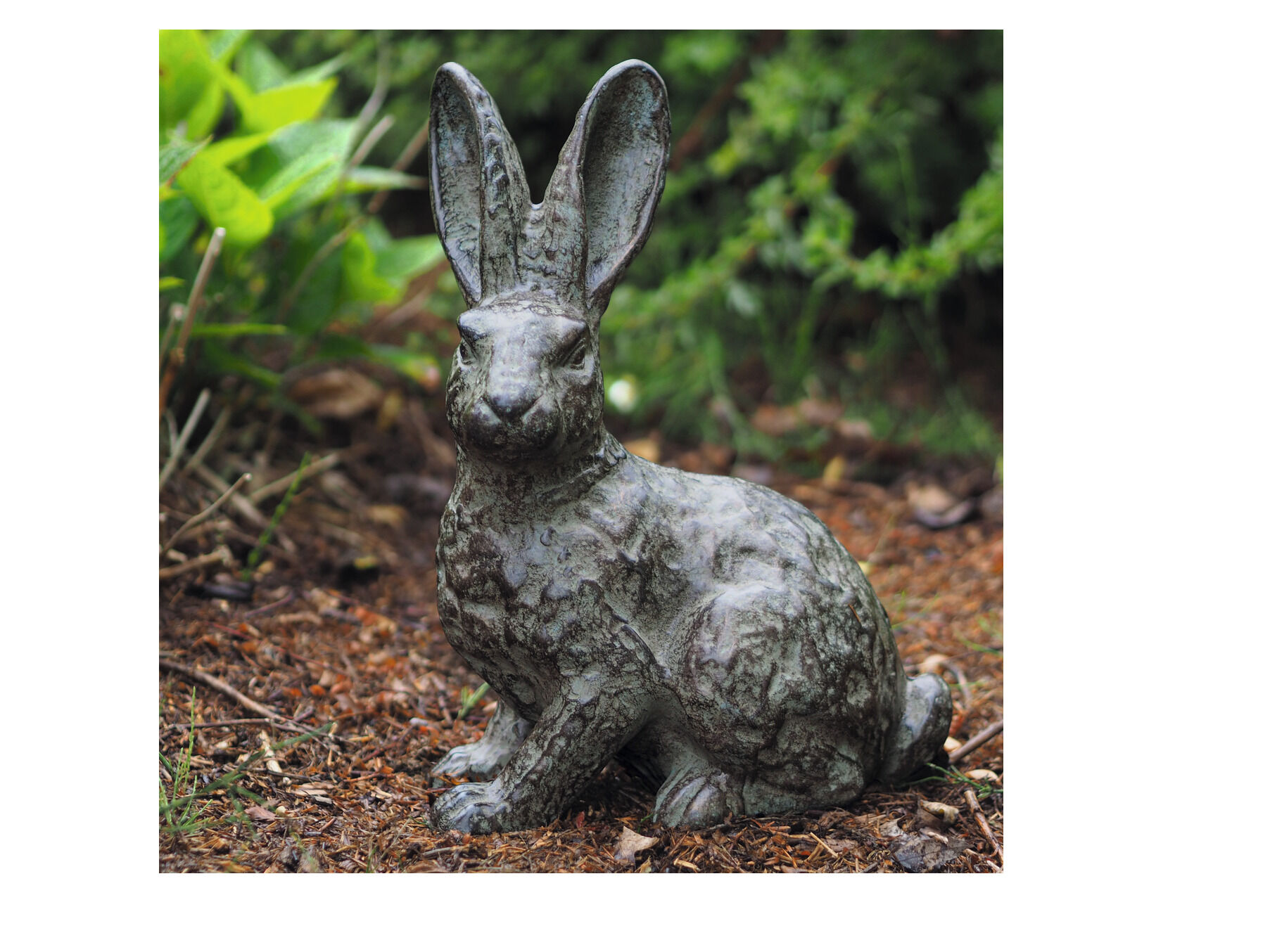 Eine bronzene Gartenskulptur in Form eines Hasen, umgeben von grünen Pflanzen und braunem Waldboden.