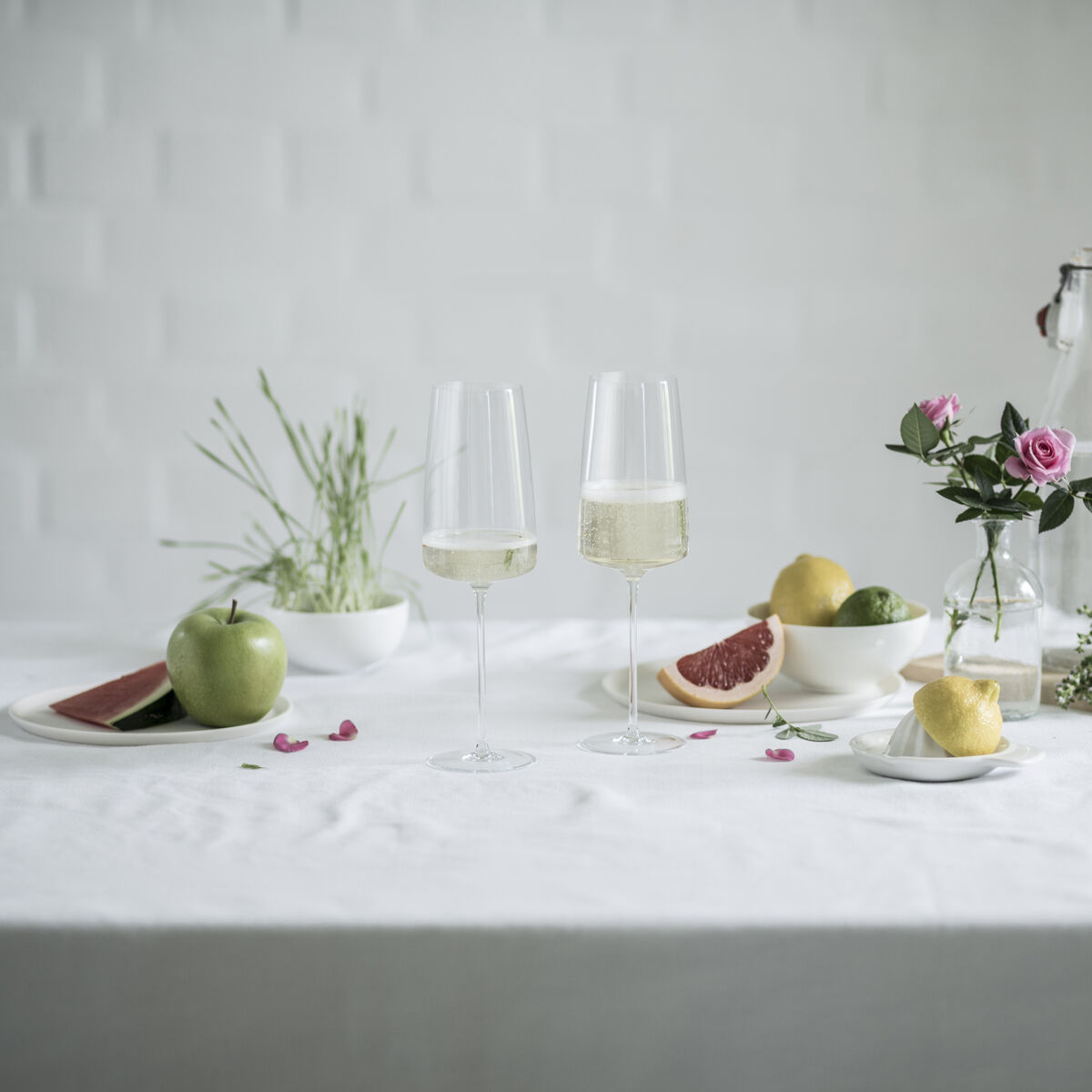 Ein Set von zwei eleganten Sektgläsern mit Inhalt steht auf einem Tisch. Der Tisch ist dekoriert mit verschiedenen Früchten, Blumen und Kräutern. Im Hintergrund ist ein helles, minimalistisches Design zu sehen.