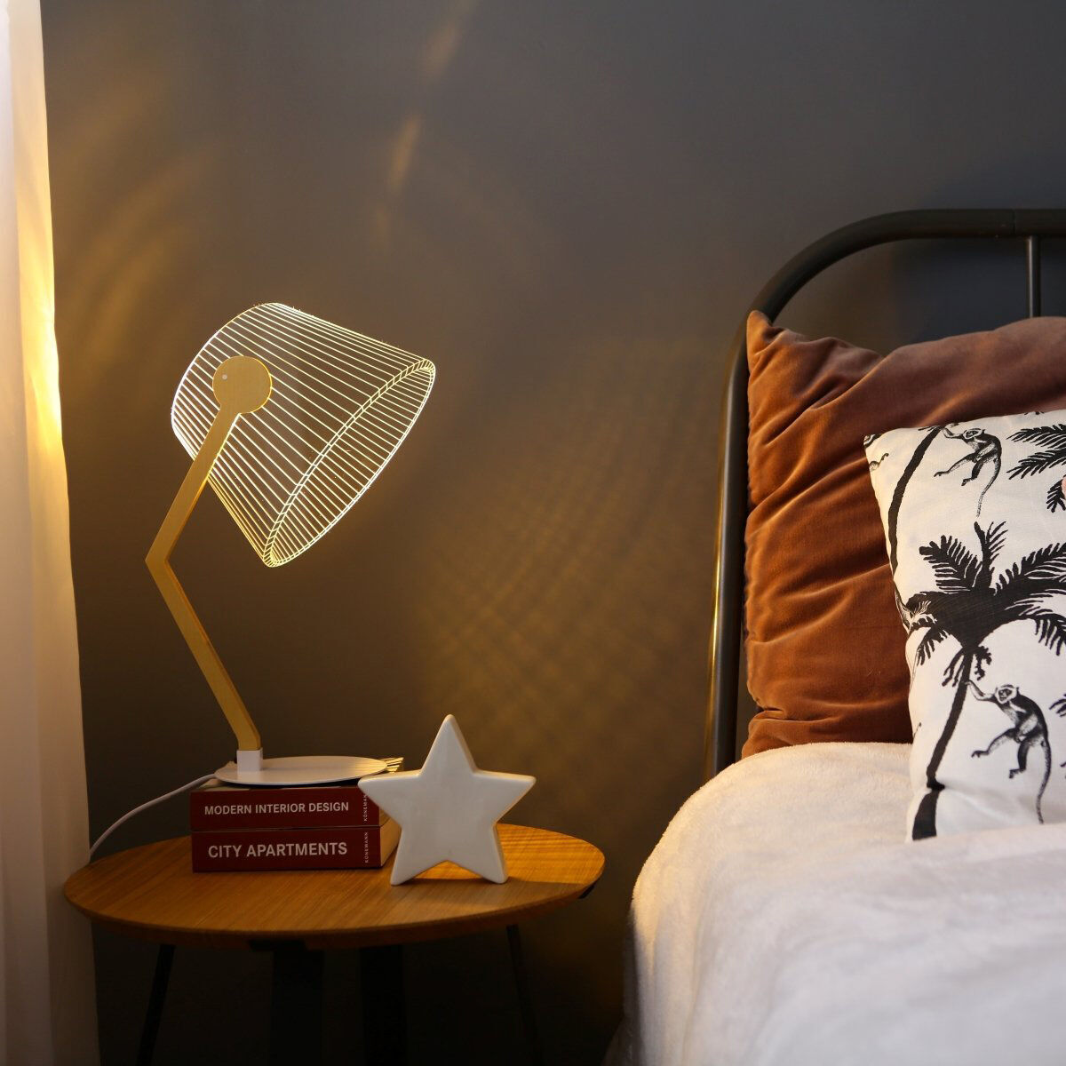Eine 3D-Lampe mit einem ZigZag-Design steht auf einem runden Holztisch neben einem Bett mit Zierkissen und weichen Decken. Im Hintergrund ist eine graue Wand zu sehen, die mit sanftem Licht beleuchtet wird.