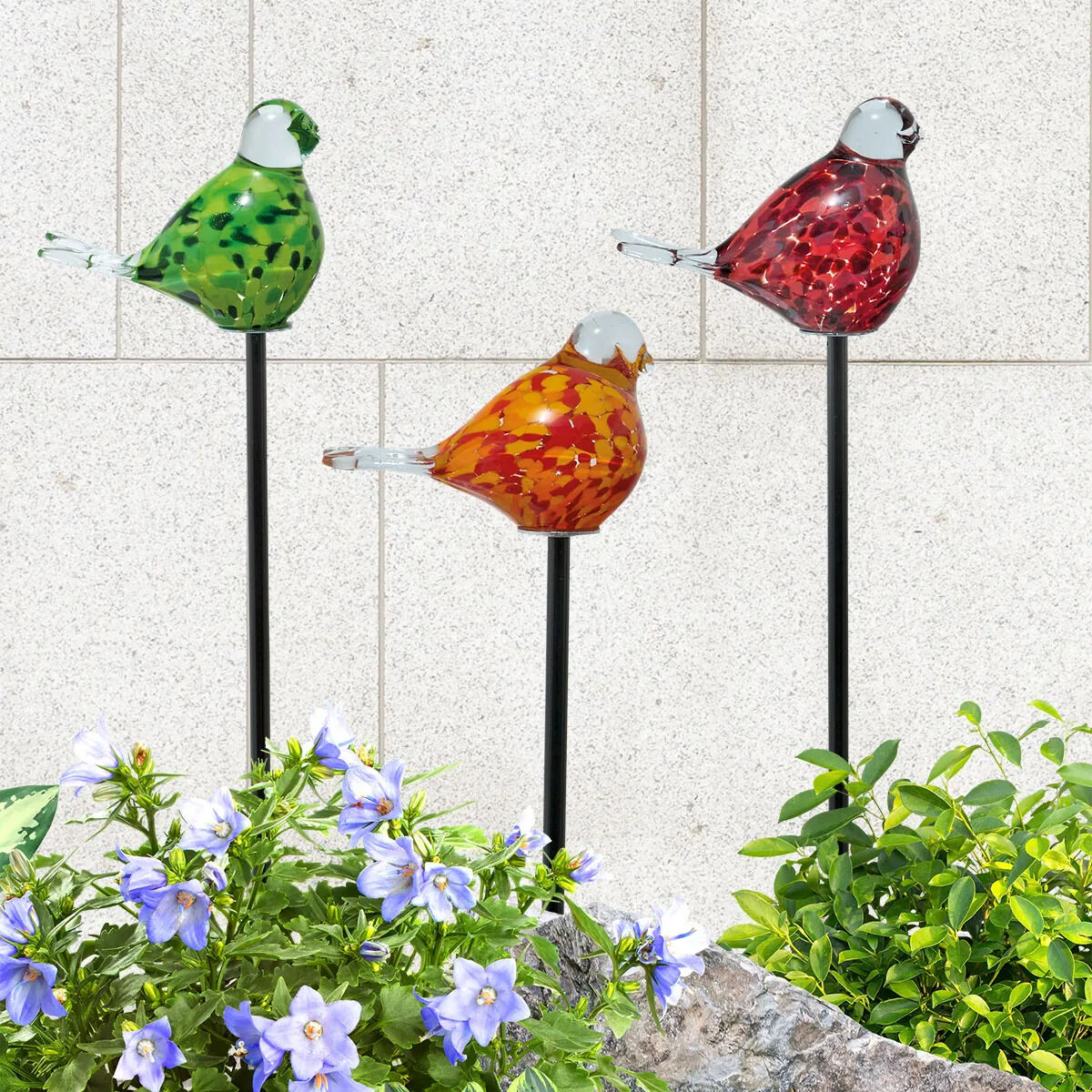 Drei bunt gesprenkelte Gartenstecker in Form von Vögeln aus Glas und Metall stehen vor einer hellen Steinwand. Einer ist grün, der zweite orange, der dritte rot. Vorne im Bild sind blühende, lilafarbene Blumen und grüne Pflanzen sichtbar.