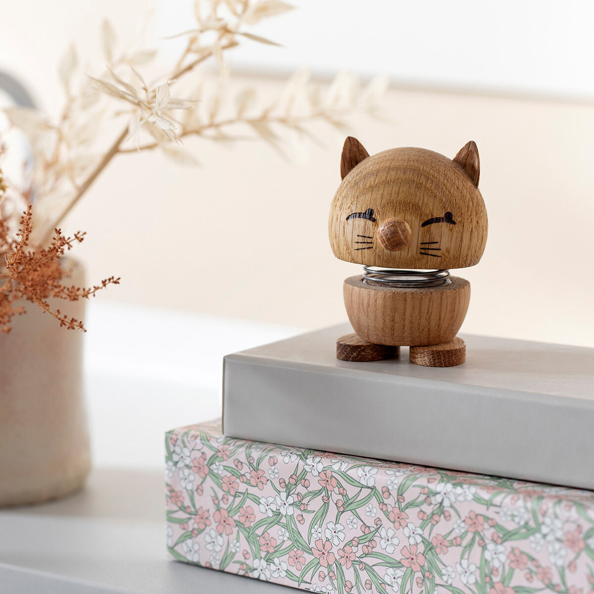 Ein skandinavisches Designobjekt in Form einer Katze aus Eichenholz, platziert auf einem dekorativen Geschenkpapier mit floralen Mustern. Die Figur hat niedliche Ohren und einen freundlichen Ausdruck, perfekt für Katzenliebhaber und als stilvolle Wohn dekoration.
