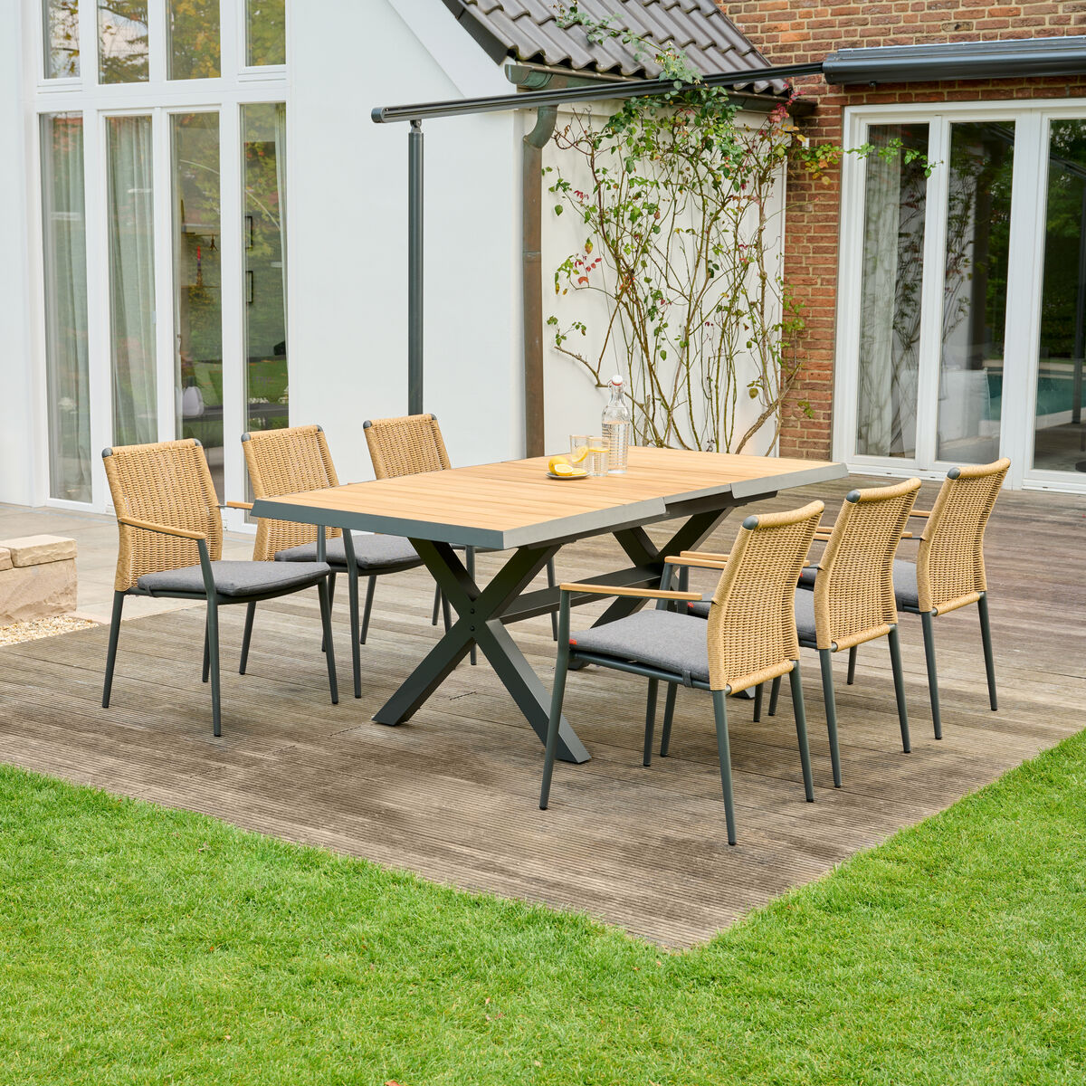 Ausziehtisch »Sincro« auf einer Terrasse mit sechs Rattan-Stühlen. Der Tisch hat eine Holzplatte mit einem robusten Metallgestell. Umgeben von einem grünen Rasen und einer modernen Hausfassade.