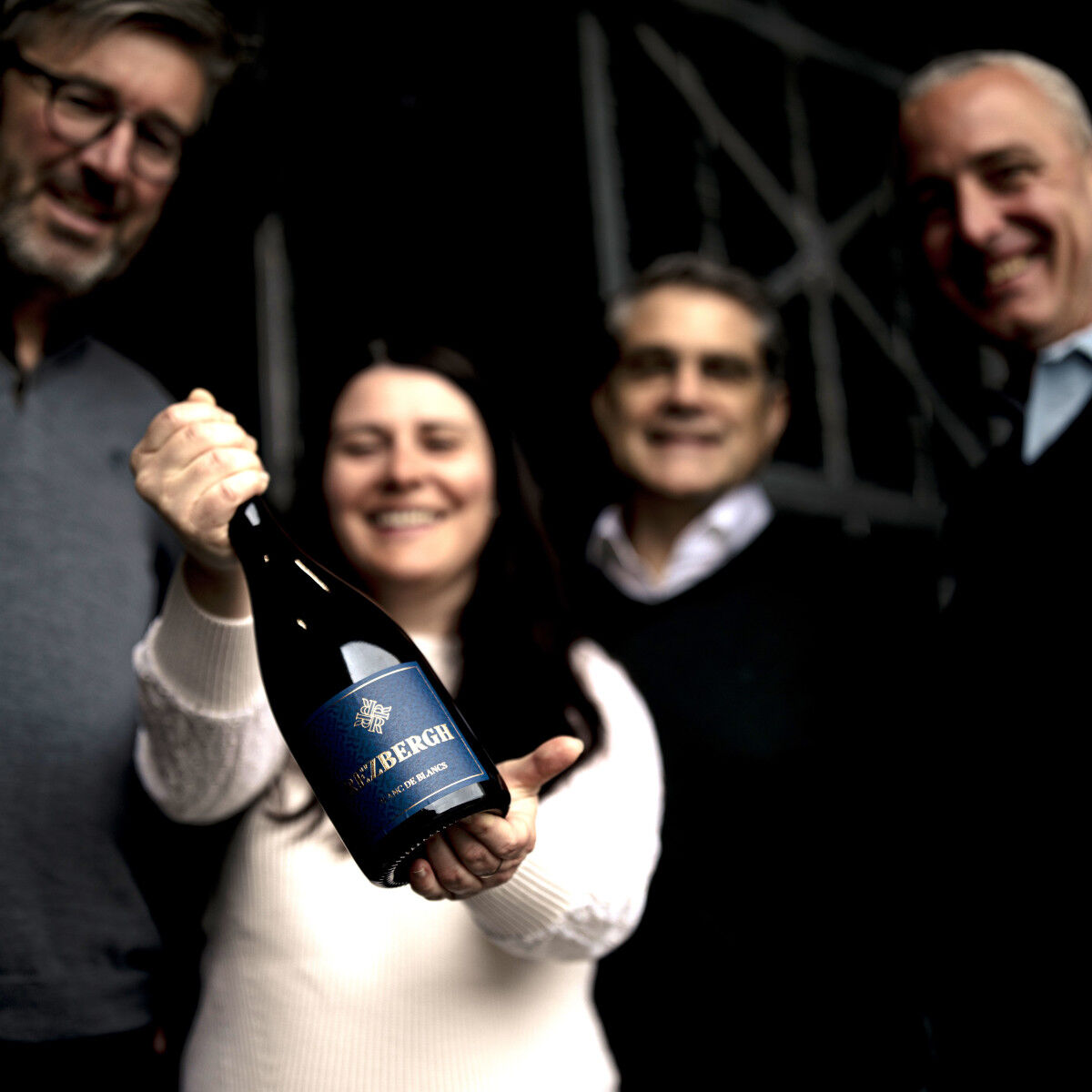 Eine Frau hält eine Flasche Crémant de Luxembourg Brut Blanc des Blancs Réserve in die Kamera, umgeben von drei lachenden Erwachsenen in eleganter Freizeitkleidung vor dunklem, unscharfem Hintergrund. Geeignet für festliche Anlässe und Feiern.