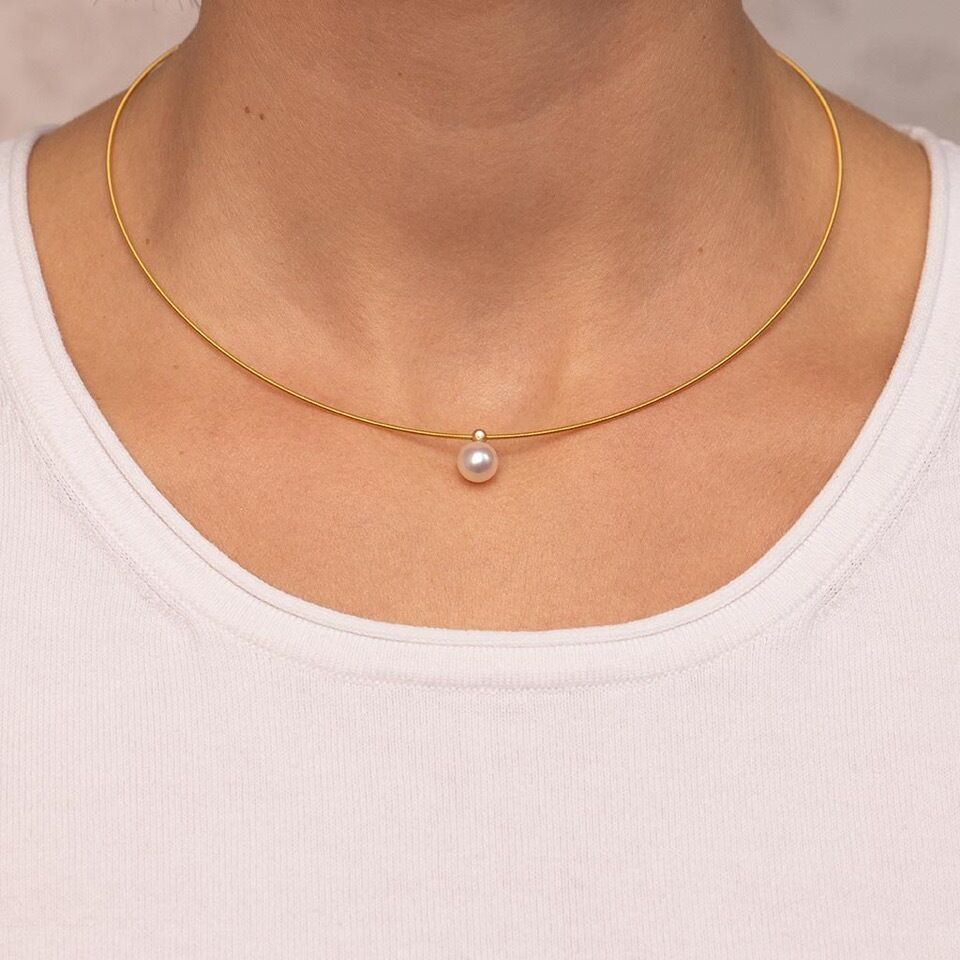 Naheaufnahme eines eleganten Colliers namens Collier Duo: Eine feine, goldfarbene Halskette mit einer mittig platzierten, runden, weißen Perle. Das Collier liegt an einem Hals, kombiniert mit einem weißen, schlichten Shirt.