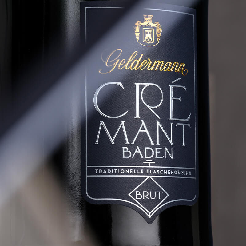 Eine Flasche Crémant Geldermann Brut mit traditioneller Flaschengärung aus Baden, versehen mit einem eleganten schwarzen Etikett und goldener Schrift, präsentiert inklusive schützender Neoprenmanschetten.