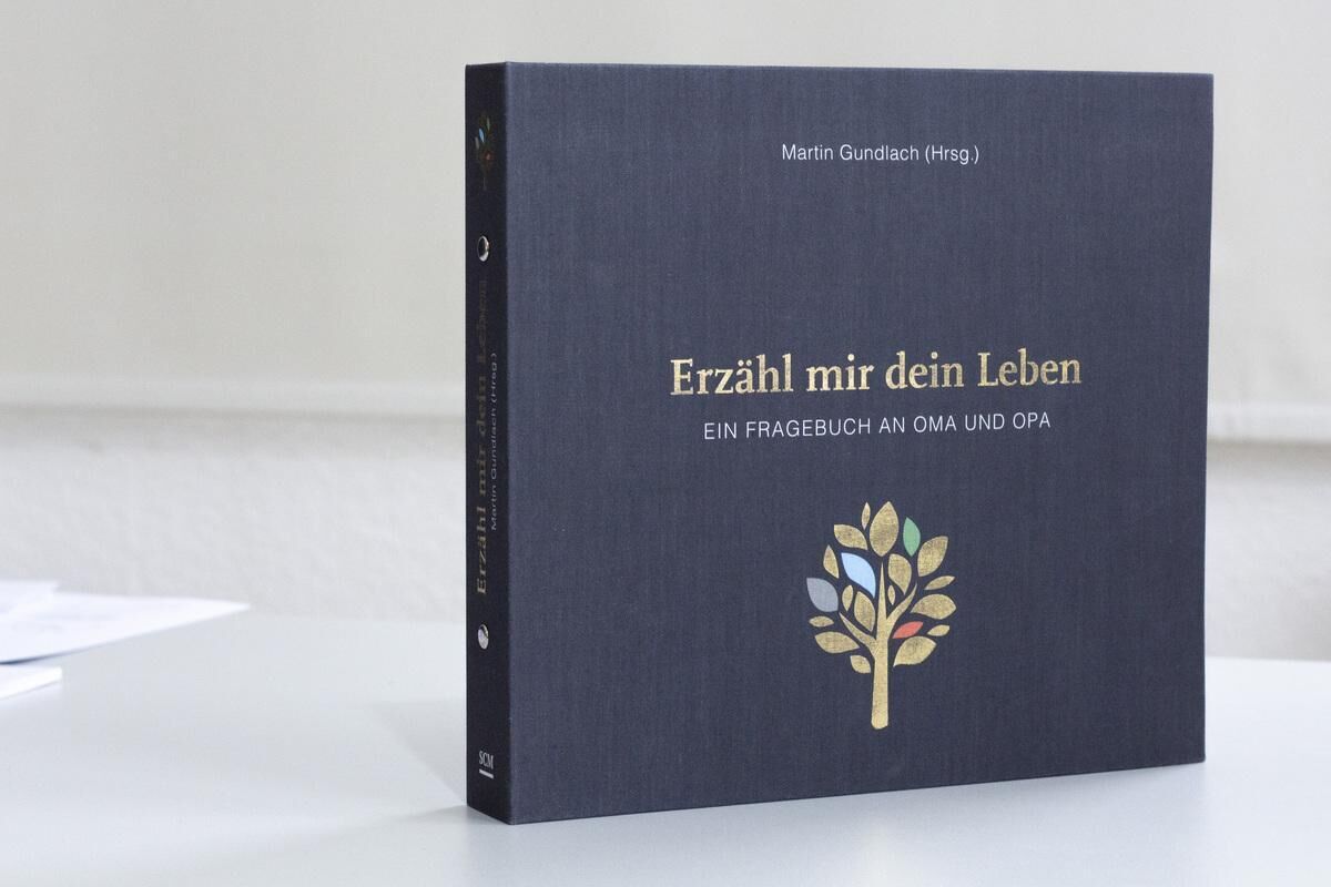 Das Bild zeigt das Erinnerungsbuch 'Erzähl mir dein Leben - Leinenausgabe'. Es hat einen schwarzen Einband mit goldenem Text und einem farbigen Baum auf dem Cover. Das Buch dient als Fragebuch an Oma und Opa, um Lebensgeschichten aufzuschreiben.