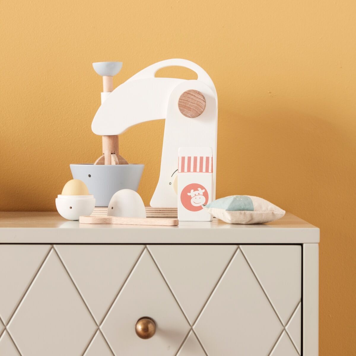 Ein Mixer-Set aus Holz mit Zubehör steht auf einer grau-gemusterten Kommode vor einer gelben Wand. Das Set umfasst einen weißen Holzmixer, einen Becher, ein Ei und weitere Accessoires.
