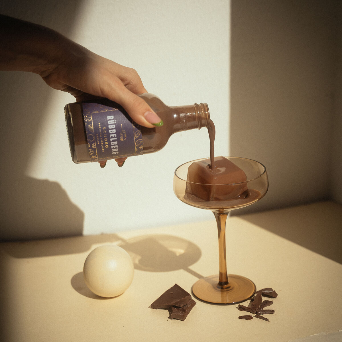 Eine braune Flasche Premium Eierlikör wird in ein edles Cocktailglas gegossen. Im Hintergrund liegen eine weiße Kugel und Stücke dunkler Schokolade auf einer hellen Oberfläche. Die Szene ist in weiches Licht getaucht und vermittelt einen luxuriösen, genussvollen Moment.