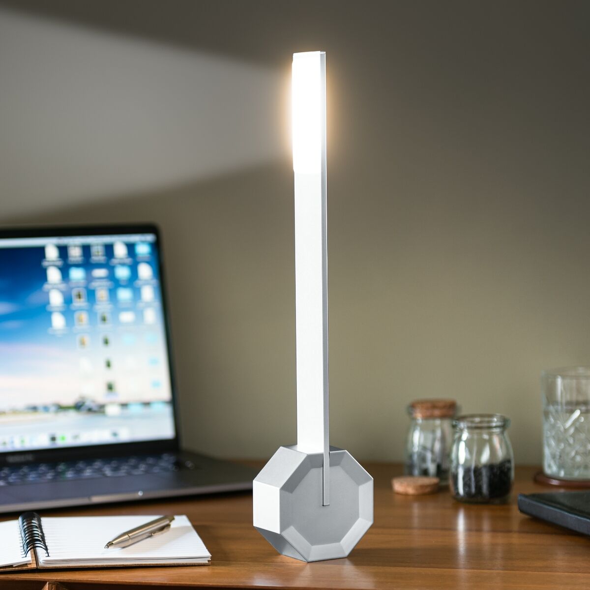 Eine moderne Octagon Tischlampe »One« steht auf einem Schreibtisch neben einem Notebook, Notizbuch und zwei Gläsern. Die Lampe hat ein minimalistisches Design und strahlt ein dezentes Licht aus. Hintergrund ist ein Teil einer Wand.
