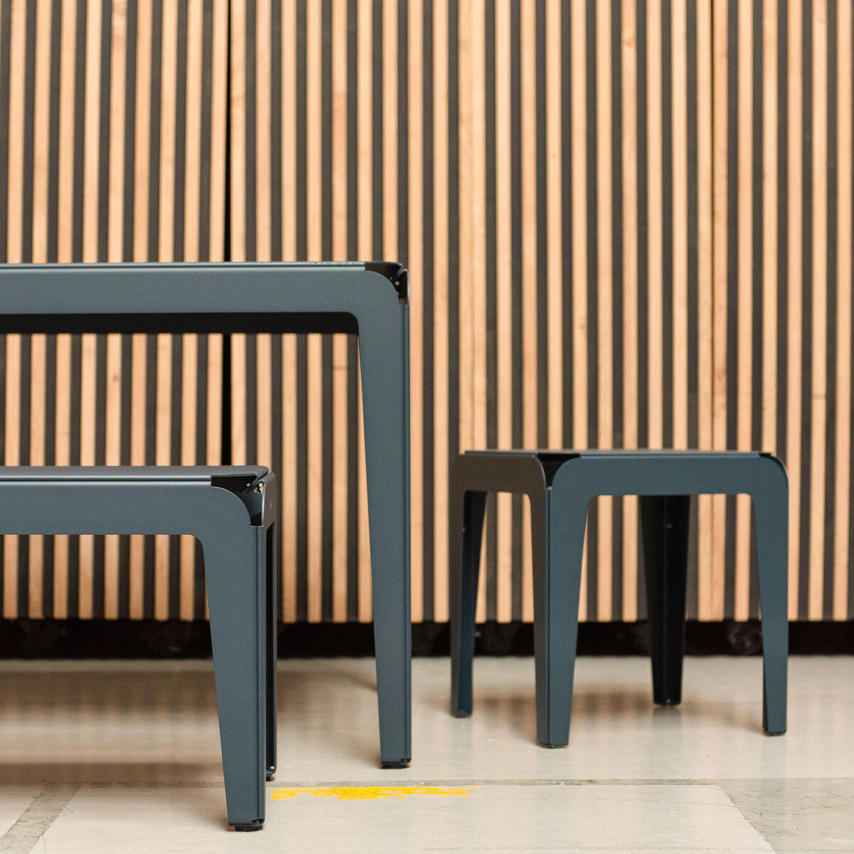 Ein moderner Hocker namens 'Bended Stool' aus Metall, abgebildet vor einer Holzwand in einem minimalistischen Design.
