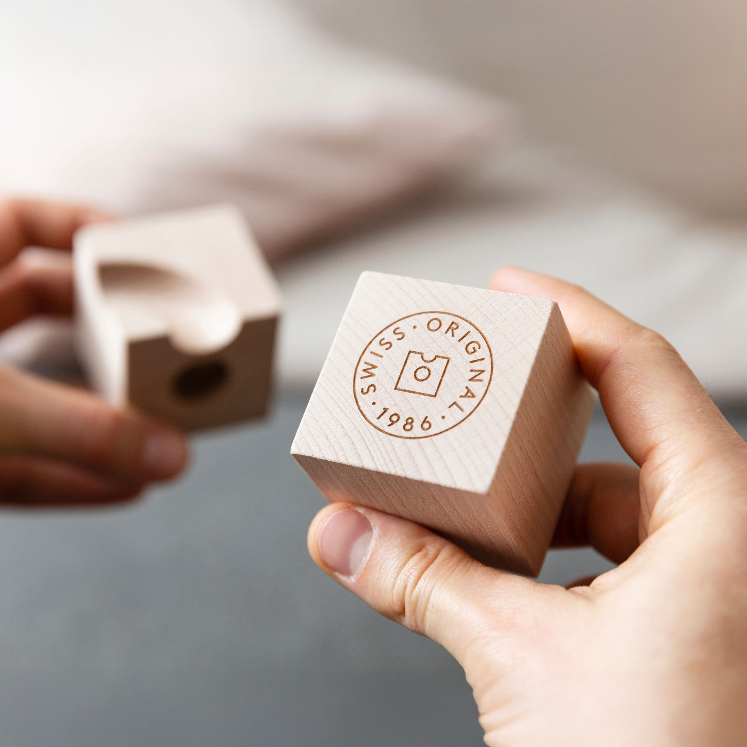 Zwei Hände halten naturbelassene, helle Holzwürfel mit präzise gefrästen Bahnteilen eines Kugelbahnsets von Cuboro. Einer der Würfel zeigt das Schweizer Qualitätssiegel. Die Szene wirkt freundlich, modern und klar fokussiert, ideal für Kinder ab 3 Jahren.