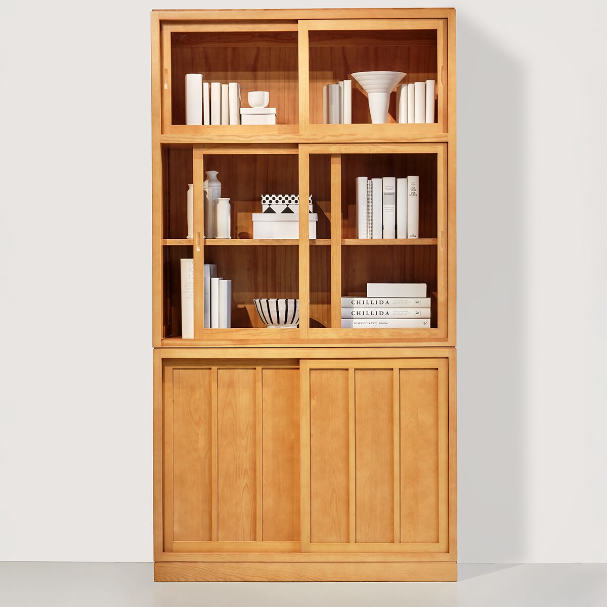 Vitrine 'Multimodus' von MARKTEX aus honigfarben lasierter spanischer Pinie, stehend vor weißer Wand. Sechs Schiebetüren oben mit Glas- und Holzfüllungen, unten massive Holzpaneele. Innen Bücher, Keramikschalen, Deko-Objekte. Zeitloses, minimalistisches Design, hochwertige Verarbeitung.