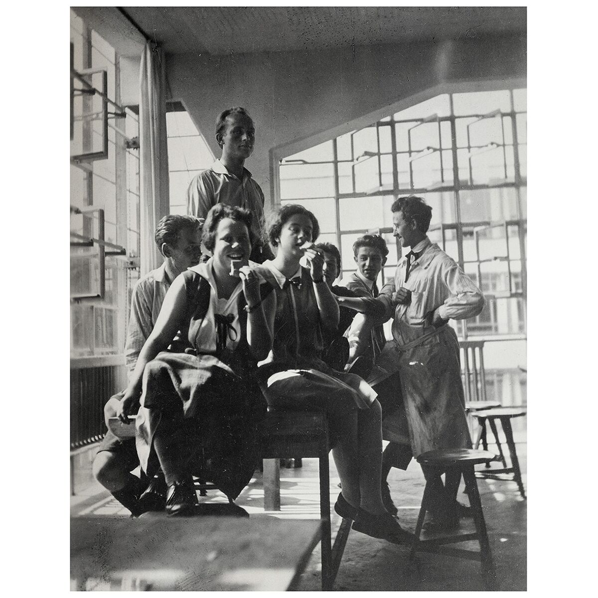 Schwarzweißfotografie einer Gruppe junger Menschen in einem hellen Atelier, vermutlich in den 1920er Jahren am Bauhaus Archiv Berlin, mit großen Fenstern im Hintergrund, typisch für das moderne Bauhaus-Design.