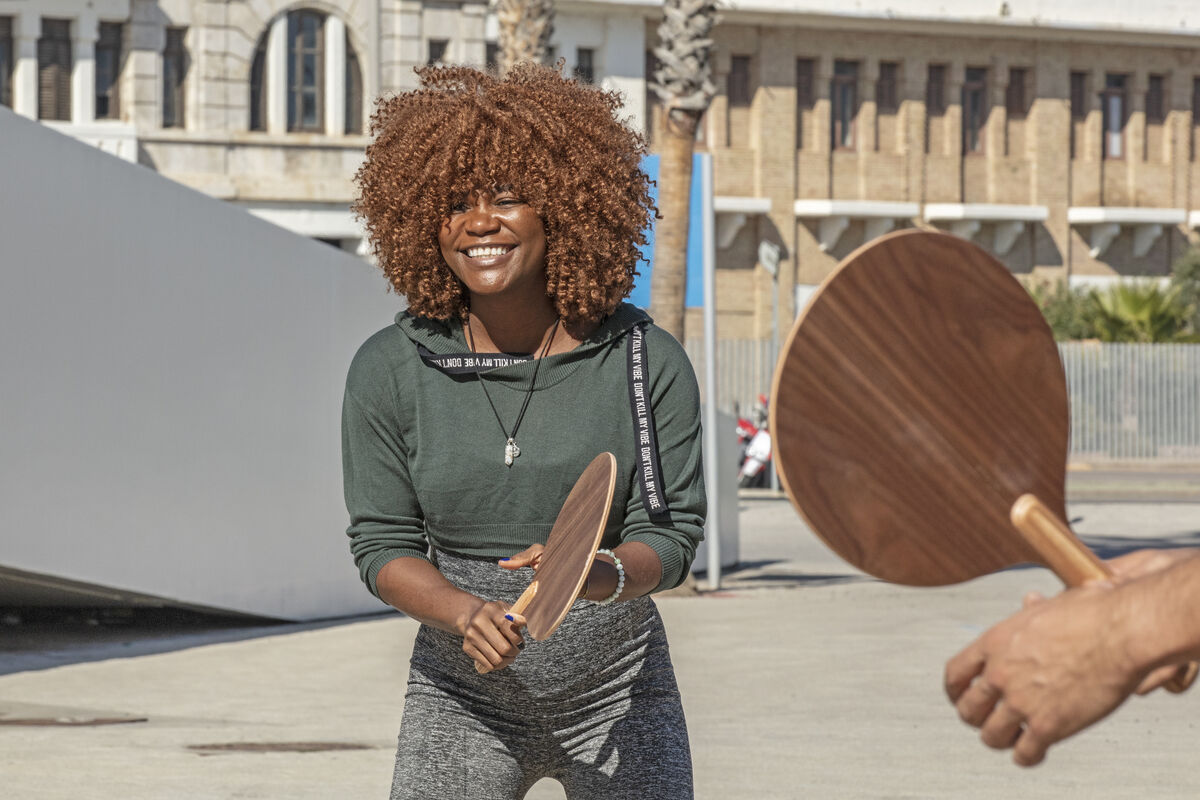Mehrere Menschen spielen unter freiem Himmel mit einem Beachball-Set. Sie lächeln und haben Spaß. Das Wetter ist sonnig, sie tragen bequeme Freizeitkleidung. Im Hintergrund ist ein Gebäude und eine Palme zu sehen.