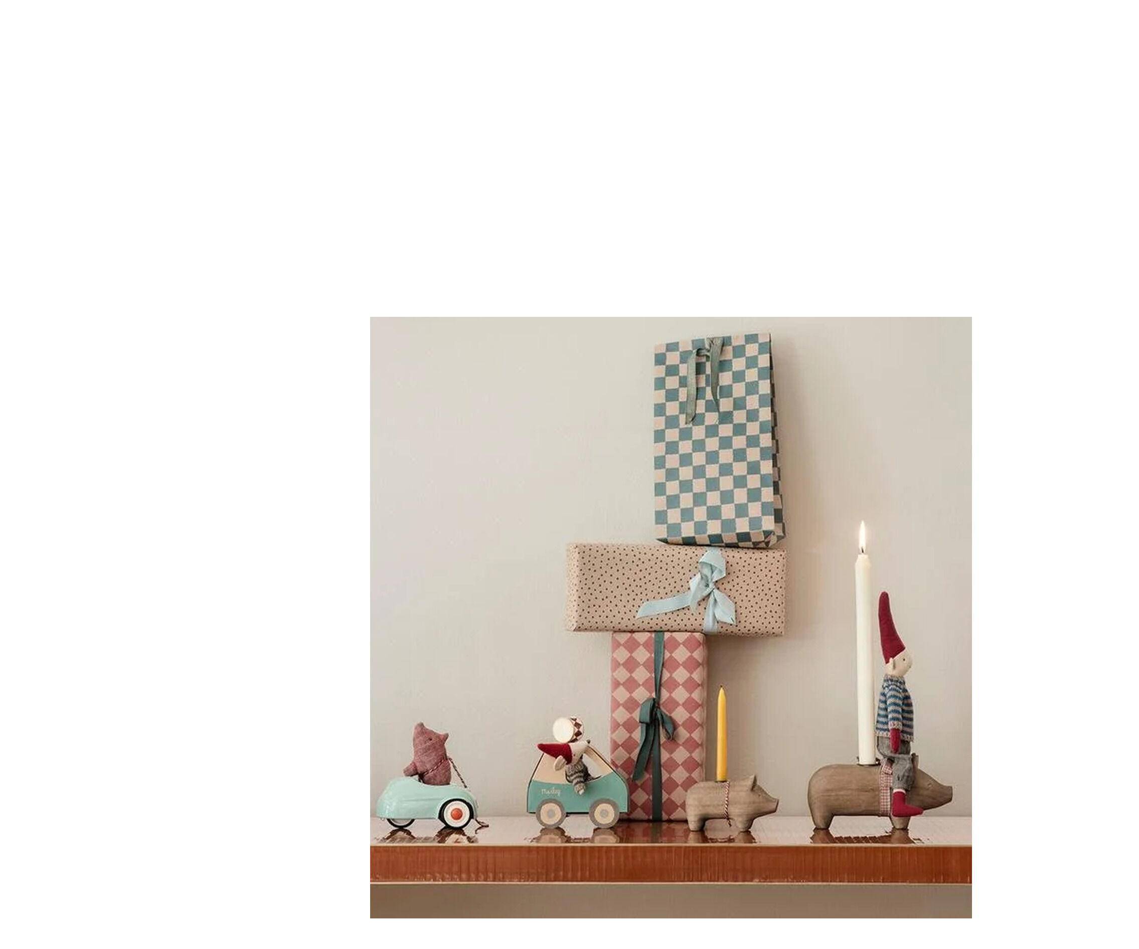 Auf einem Regal stehen mehrere Maileg-Figuren, darunter die Weihnachtsmaus im grünen Auto mit roter Zipfelmütze und Strickpullover, umgeben von Tieren mit Kerzen und weihnachtlicher Deko. Darüber hängen festliche Girlanden; im Hintergrund bunte Geschenke.