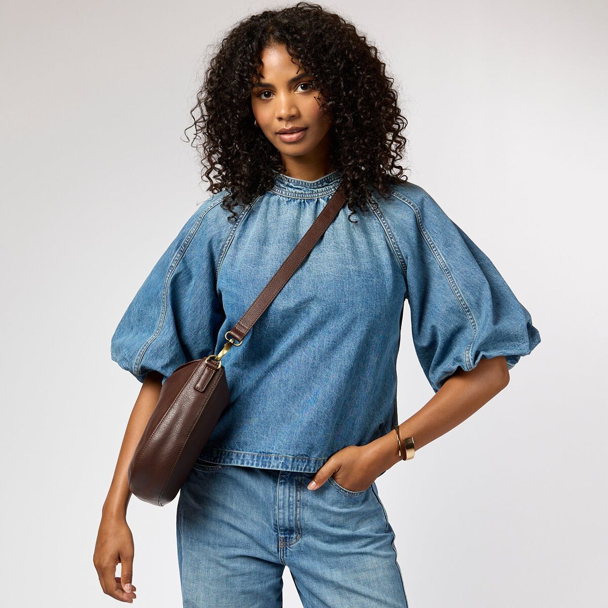 Frau trägt eine dunkelbraune Umhängetasche »Lana« zu einem Jeansoutfit mit langärmliger Bluse und steht vor neutralem Hintergrund. Die Tasche ist aus Leder, hat goldene Details und einen verstellbaren Riemen.