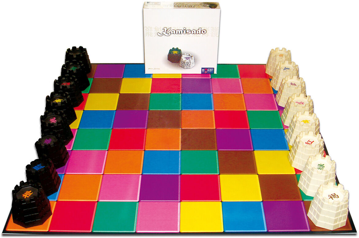 Das Bild zeigt das Brettspiel 'Kamisado'. Es besteht aus einem farbenfrohen quadratischen Brett mit unterschiedlich gefärbten Spielfeldern. An den beiden Seiten des Bretts befinden sich schwarze und weiße Spielsteine. Im Hintergrund ist die Verpackung des Spiels zu sehen.