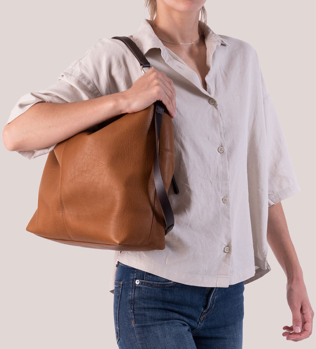 Eine Frau trägt eine braune Leder-Beuteltasche namens »Chaza Pouch Upend« über der Schulter. Sie trägt ein beigefarbenes Hemd und blaue Jeans.