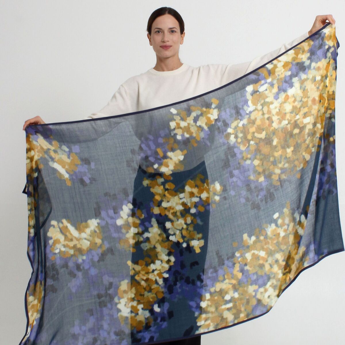 Eine Frau hält einen großen, transparenten Wollschal »Hazel« Blau der Marke Shalls Barcelona aus 100% Schurwolle ausgebreitet vor sich. Das Tuch zeigt ein florales, gelb-weiß-violettes Muster auf blauem Grund. Der Hintergrund ist neutral und schlicht.