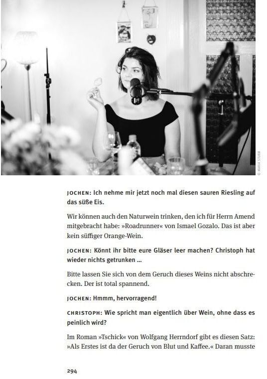 Schwarz-weiß Foto einer Frau, die an einem Tisch sitzt und in ein Mikrofon spricht. Der Tisch hat eine Lampe und Gläser darauf. Textpassagen aus dem Buch 'Alles Gesagt? Buch zum Podcast' sind auf der rechten Seite zu sehen. Im Hintergrund sind unscharfe Menschen und Gegenstände zu erkennen. Am unteren Rand der Seite steht die Zahl 294.