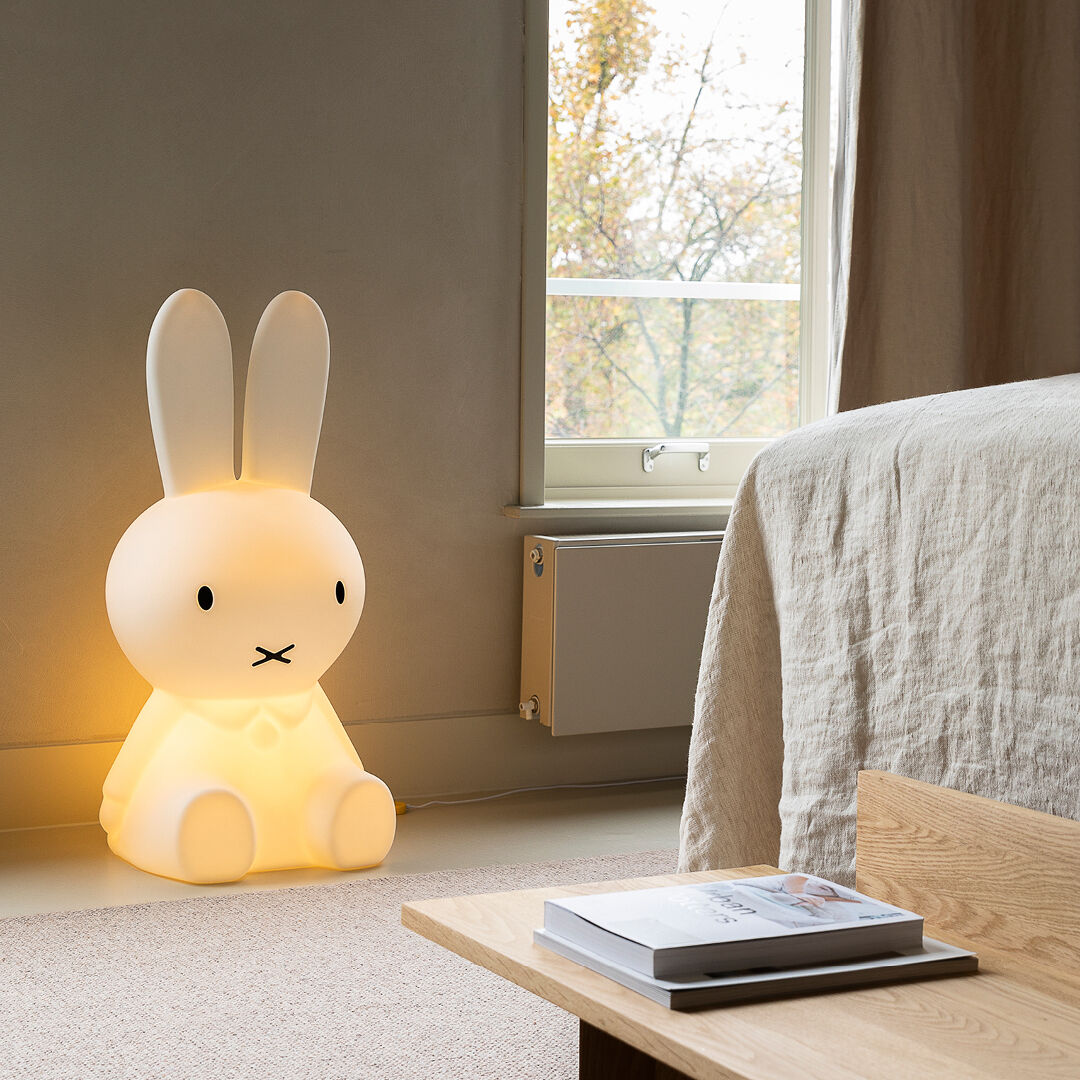 Kinderlicht »Miffy XL« in Form eines sitzenden, beleuchteten Hasen. Es steht in einem gemütlichen Schlafzimmer neben einem Bett und erhellt den Raum mit einem sanften gelben Licht. Ein Fenster im Hintergrund zeigt eine herbstliche Szene.
