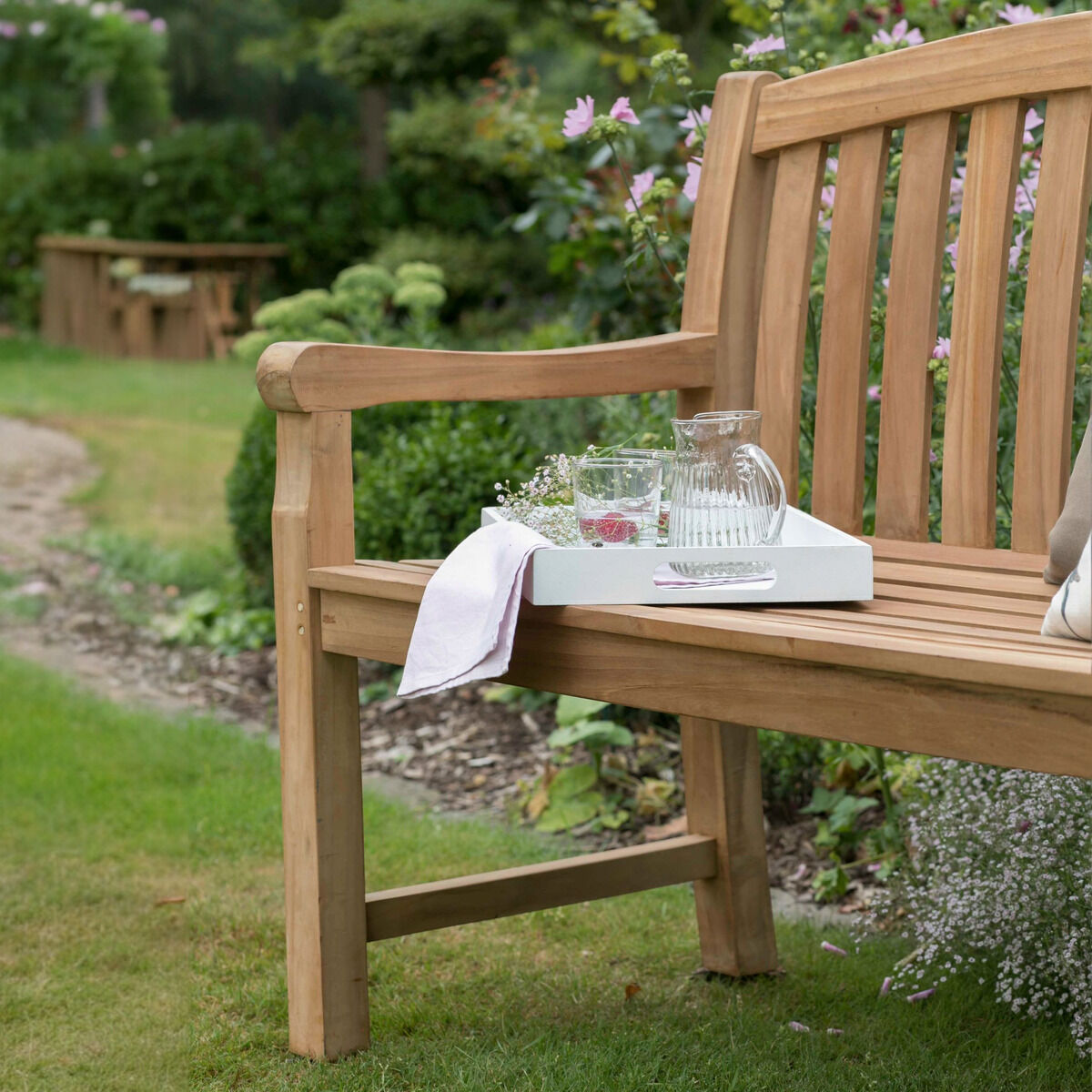 Nahansicht der unbehandelten Teak-Gartenbank »Swindon« von bene living in einem üppig bepflanzten Garten. Die Bank hat breite Armlehnen und eine hohe, leicht geschwungene Rückenlehne aus senkrechten Holzlatten. Auf dem Sitz stehen Tablett, Glaskaraffe und zwei Gläser; ein weißes Tuch hängt über der Armlehne. Im Hintergrund verläuft ein Kiesweg zu einer weiteren Bank.