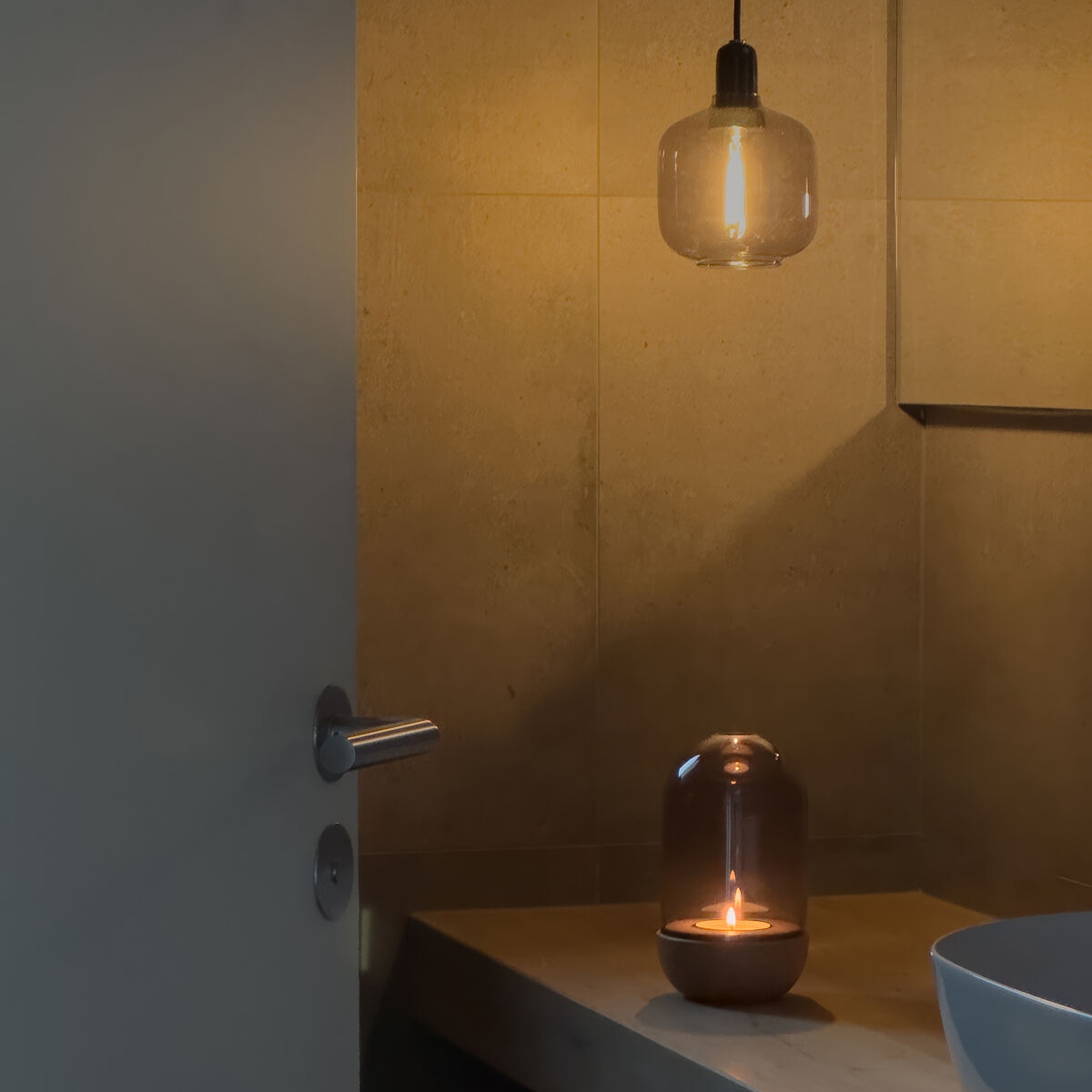 Im stimmungsvoll beleuchteten Badezimmer steht auf einer hellen Ablage das Windlicht »GLOW« von finbeaux mit Rauchglas von Christoph Schoen. Daneben hängt eine Pendelleuchte, deren warmes Licht eine gemütliche Atmosphäre erzeugt. Modernes, stilvolles Ambiente.