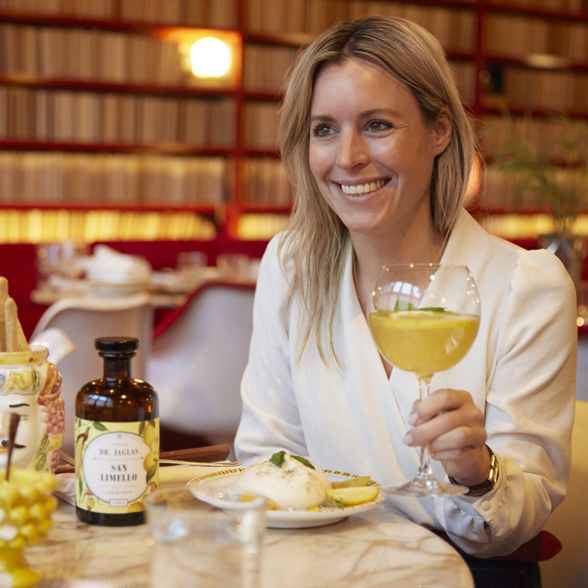 Eine Frau in weißer Bluse sitzt in einem Restaurant an einem Tisch mit einem Glas San Limello, einem alkoholfreien Aperitif. Auf dem Tisch stehen eine Flasche San Limello, ein Zitronen-Dekor und eine Schale Grissini. Der Hintergrund zeigt rote Sofas und Bücherregale.