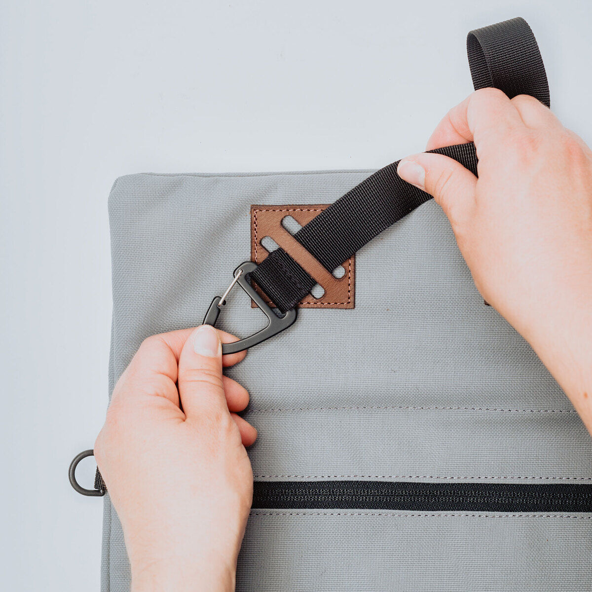 Zwei Hände, die eine graue Tasche mit einem abnehmbaren schwarzen Gurt und braunen Akzenten halten; sicherer Clip und minimalistisches Design.