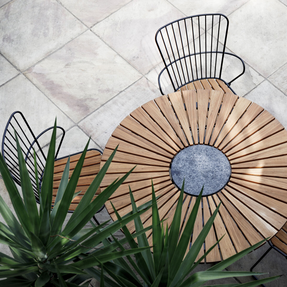 Eine Draufsicht auf vier schwarze „PAON“-Esszimmerstühle um einen runden Holztisch auf einer gefliesten Terrasse. Die Stühle haben eine Metallrahmenkonstruktion und Holzlatten-Sitzflächen. Eine grüne Pflanze ist unten links im Bild sichtbar.