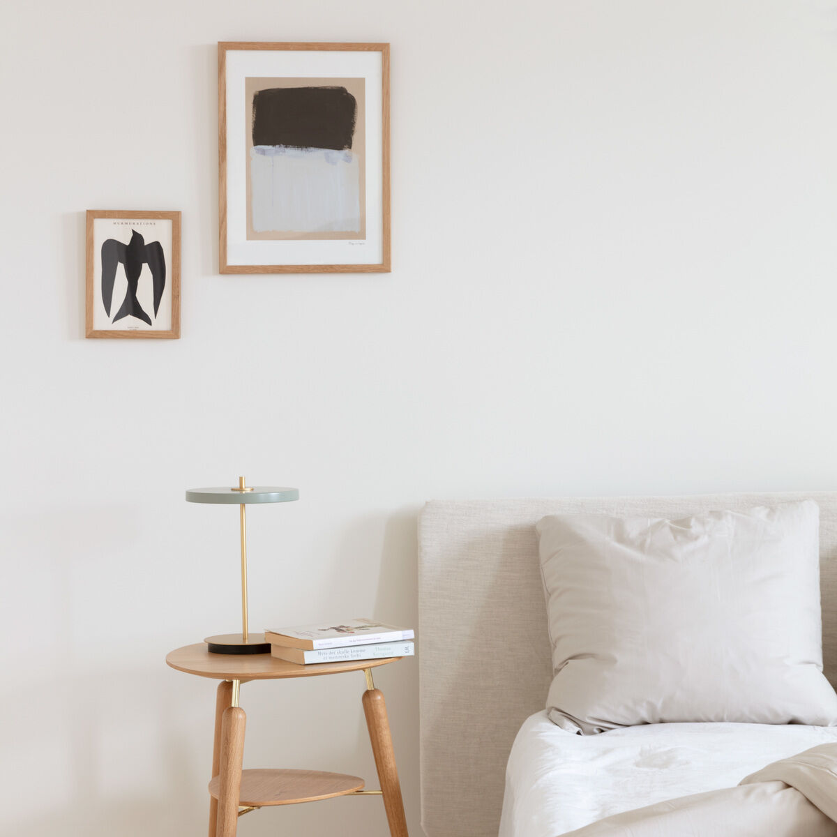 Eine moderne Tischlampe 'Asteria Move II' steht auf einem kleinen runden Holztisch neben einem Bett in einem minimalistisch eingerichteten Schlafzimmer. Die Wände sind weiß und es hängen zwei gerahmte Kunstwerke darüber. Der Raum wirkt hell und luftig, mit hölzernem Fußboden und beigen Betten.