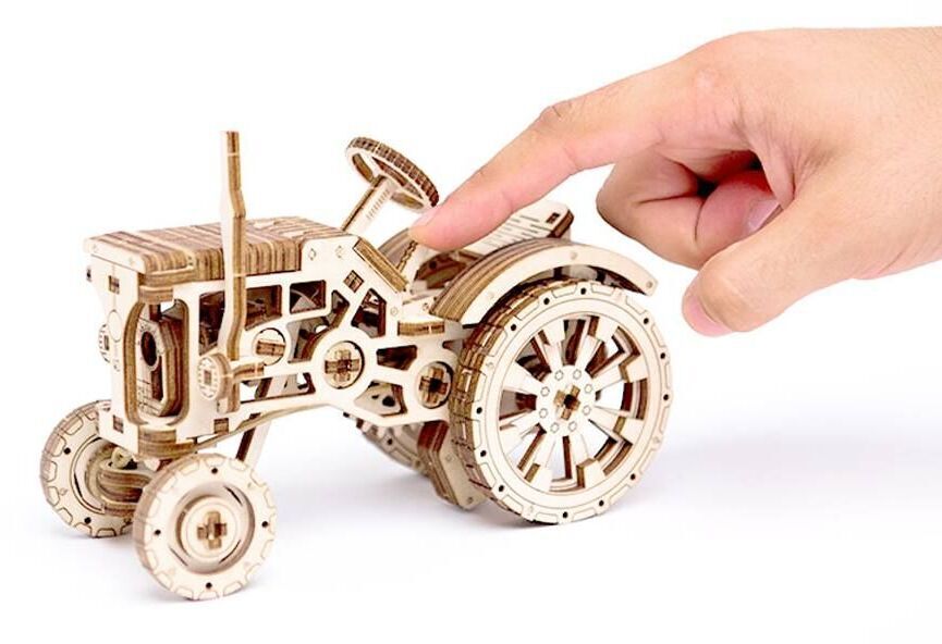 Ein 3D-Holzmodell in Form eines Traktors, das detailreich aus verschiedenen Holzelementen zusammengesetzt ist. Eine Hand zeigt auf das Modell, das komplexe mechanische Teile wie Räder und Hebel darstellt.