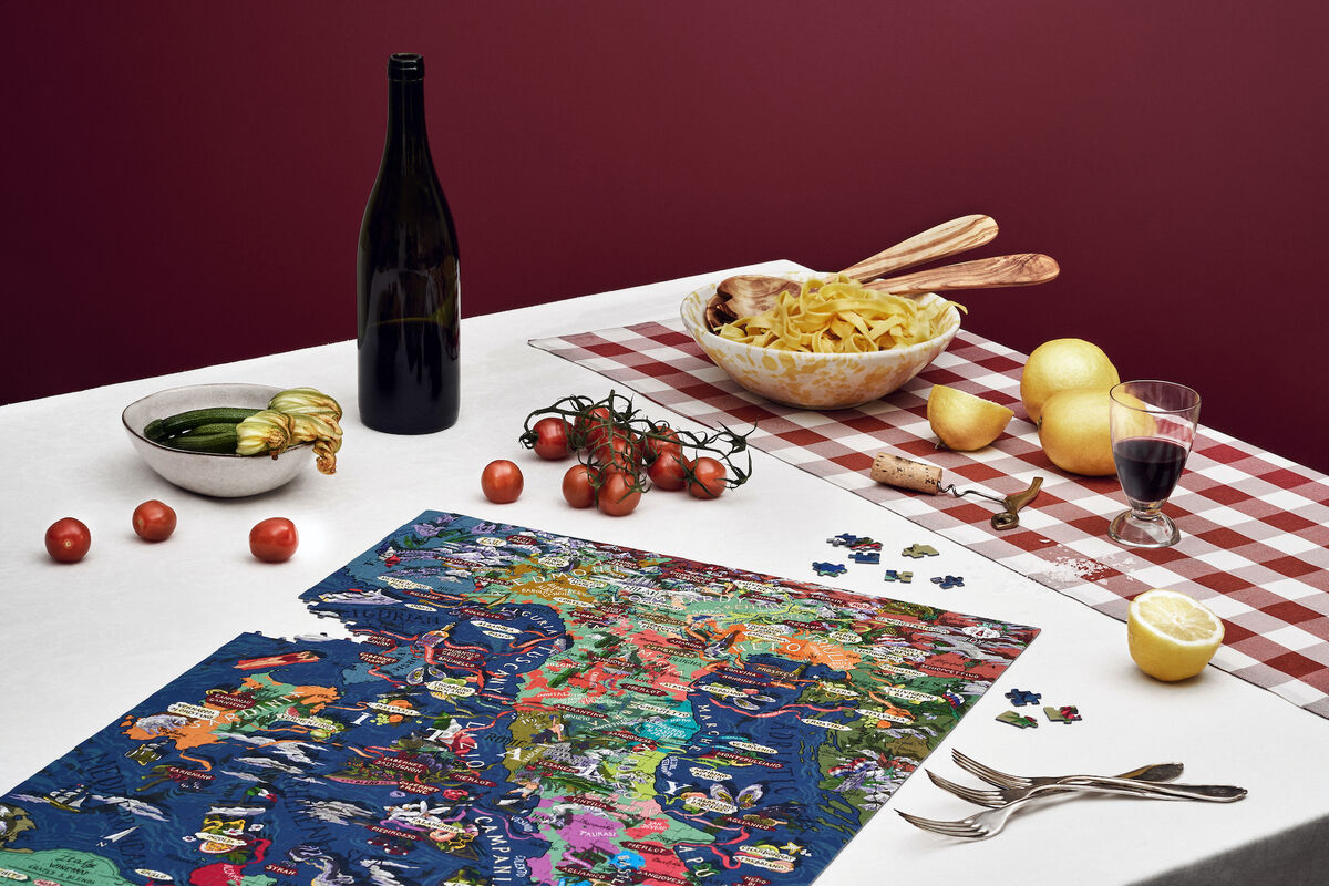 Ein Puzzle mit einer farbenfrohen Landkarte von Italien liegt teilweise zusammengesetzt auf einem Tisch. Daneben befinden sich eine Flasche Rotwein, ein Glas Wein, Tomaten, eine Zitronenhälfte, eine Schale mit Zucchini und Auberginen, ein großer Teller Spaghetti und Besteck. Der Hintergrund zeigt eine rote Wand, und der Tisch ist mit einem Weiß-Rot-karierten Tischtuch bedeckt.