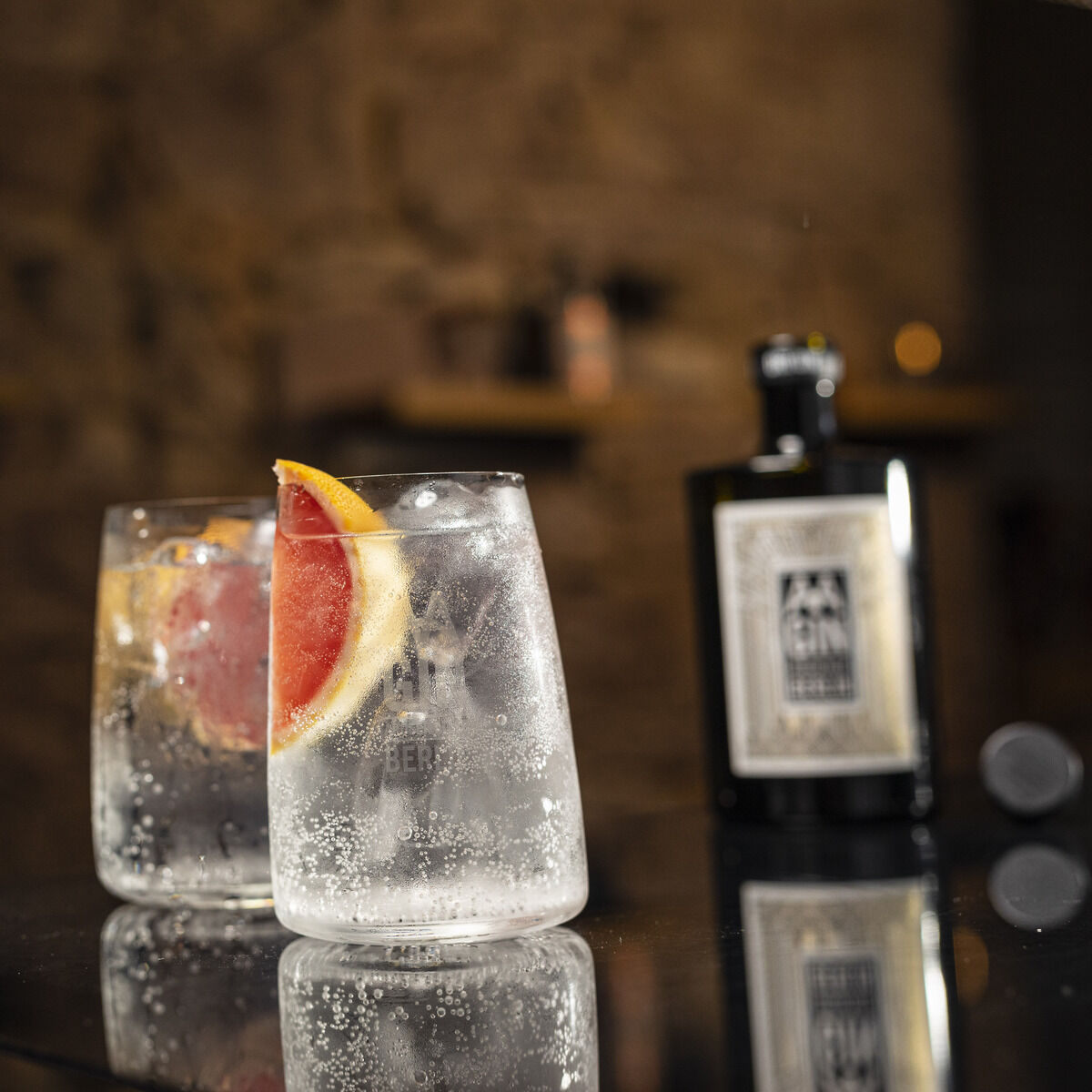 Im Vordergrund stehen zwei elegante Gläser mit Gin Tonic, Eiswürfeln und einer Scheibe Grapefruit. Dahinter eine schwarze Flasche »AAGIN« Dry Gin im Set von Weindimensional. Die Szene ist stilvoll auf reflektierendem Untergrund mit warmer, unscharfer Bar-Atmosphäre inszeniert.