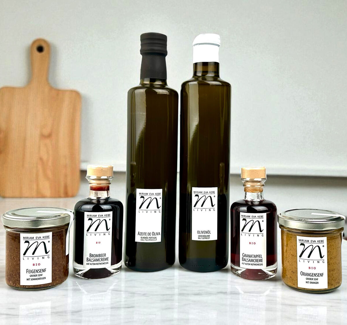 Das Bild zeigt ein Gourmet-Set bestehend aus zwei Flaschen Olivenöl und Essig sowie vier Gläsern und Fläschchen mit unterschiedlichen Feinkost-Aufstrichen und Delikatessen, arrangiert auf einer weißen Küchenarbeitsplatte vor einem Schneidebrett im Hintergrund.