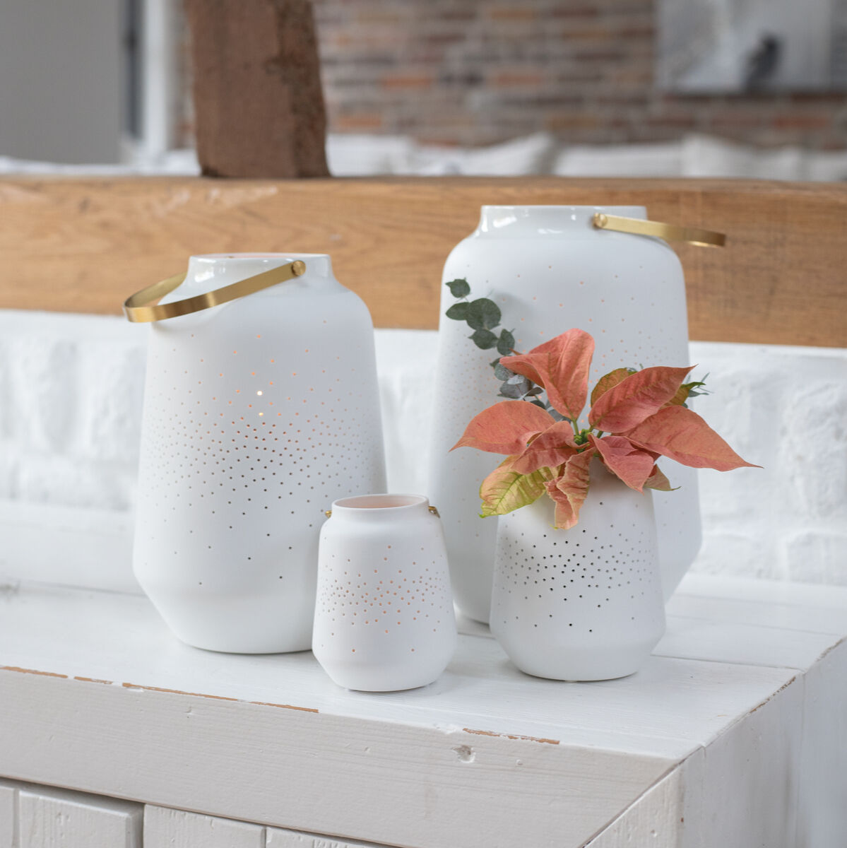 Vier weiße Porzellanlaternen in verschiedenen Größen stehen auf einer weißen Holzoberfläche. Eine Laterne ist mit goldenen Henkeln und Blumen dekoriert. Im Hintergrund sind eine Backsteinmauer und ein rustikaler Holzbalken sichtbar.
