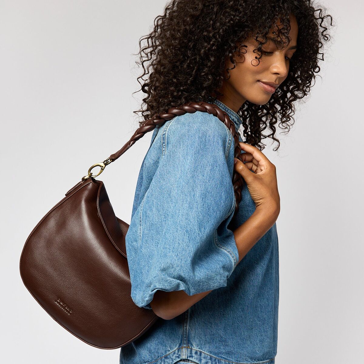 Frau trägt eine dunkelbraune Schultertasche aus Leder namens »Lana« über der Schulter. Die Tasche hat ein schlichtes, minimalistisches Design und wird zu einer blauen Jeansbluse kombiniert. Das Bild stellt die Tasche als modisches Accessoire für den Alltag dar.