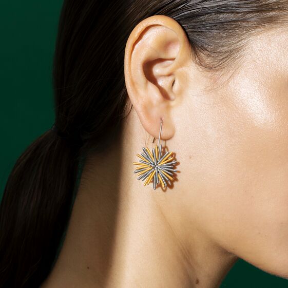 Detailaufnahme eines weiblichen Ohrs mit einem auffälligen, blumenförmigen Ohrring in Gold, Blau und Silber vor grünem Hintergrund. Das Schmuckstück wirkt modern und künstlerisch und ist für Damen geeignet.